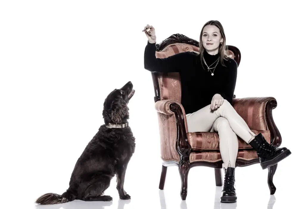 Frau sitzt auf einem luxuriösen Stuhl, während ihr Hund auf dem weißen Boden neben ihr sitzt und sie anblickt