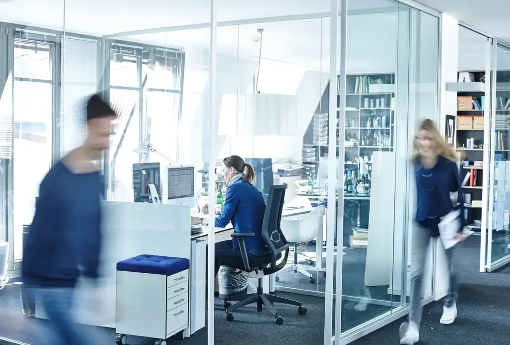 Eine Frau in einem modernen Glasbüro, von hinten gesehen, während sie konzentriert an ihrem Schreibtisch arbeitet, umgeben von transparenten Wänden