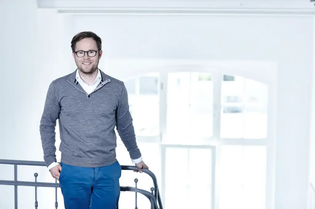 Ein Mann in grauem Oberteil und blauer Hose steht vor einem Geländer, mit Fenstern im Hintergrund