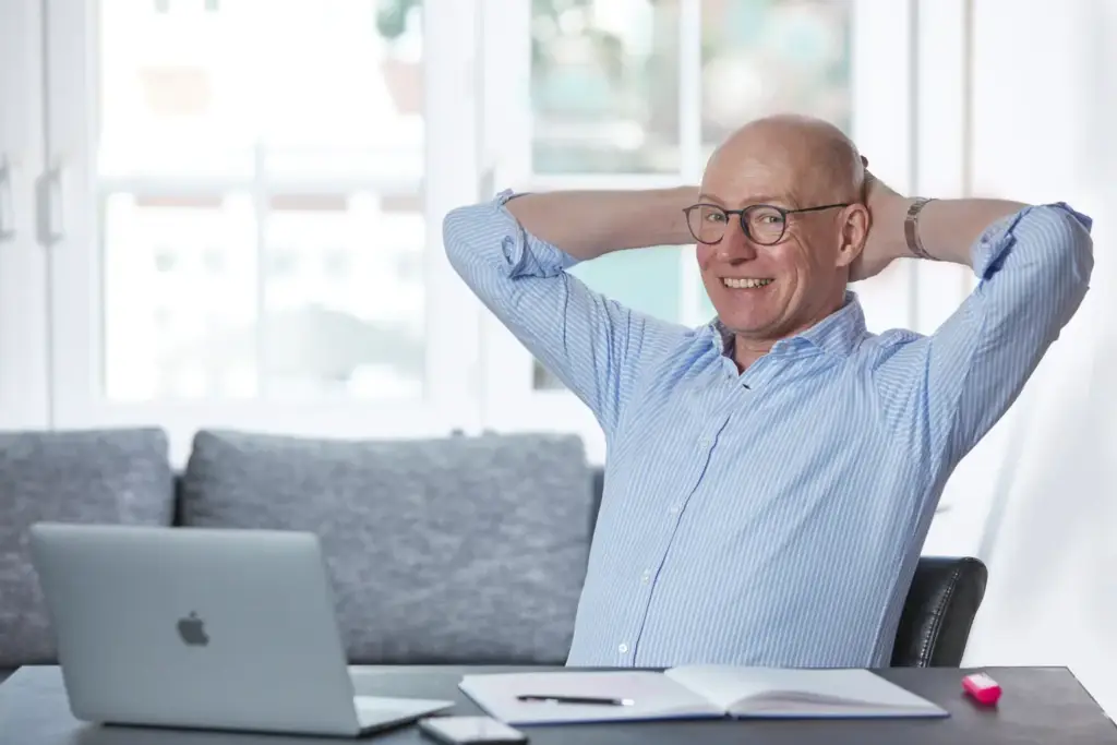 Ein Mann in einem blauen Hemd lächelt in die Kamera, während er sich entspannt auf seinem Schreibtischstuhl zurücklehnt. Auf dem Tisch stehen ein Laptop und ein Notizbuch