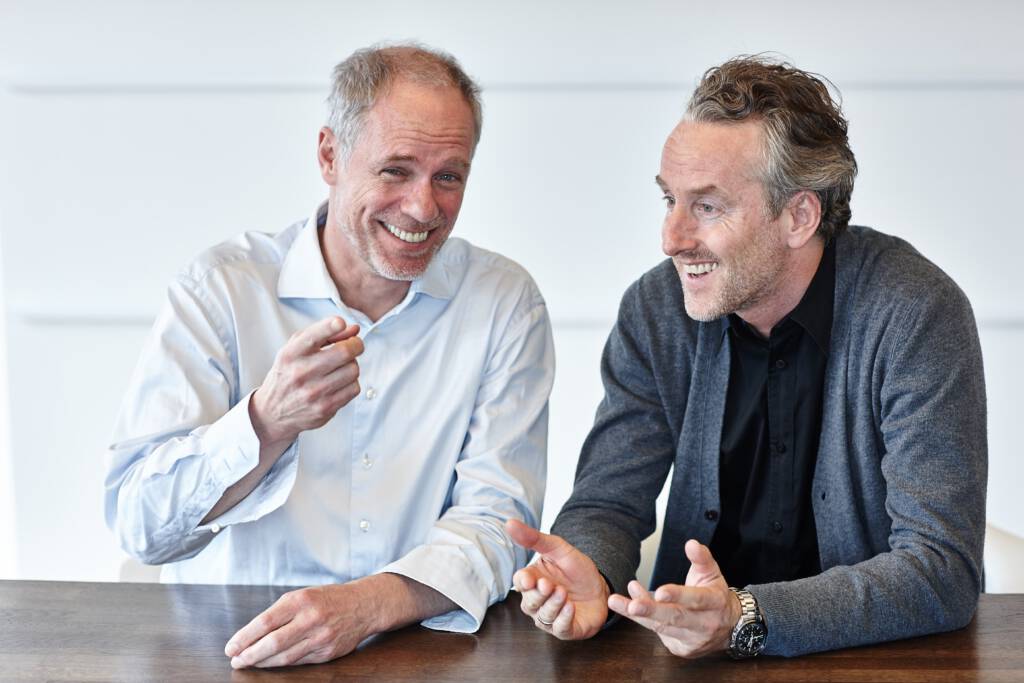 Zwei ältere, lachende Kollegen in legerer Business-Kleidung sitzen an einem Holztisch und führen ein lebhaftes Gespräch, wobei der linke Mann im blauen Hemd auf etwas zeigt.