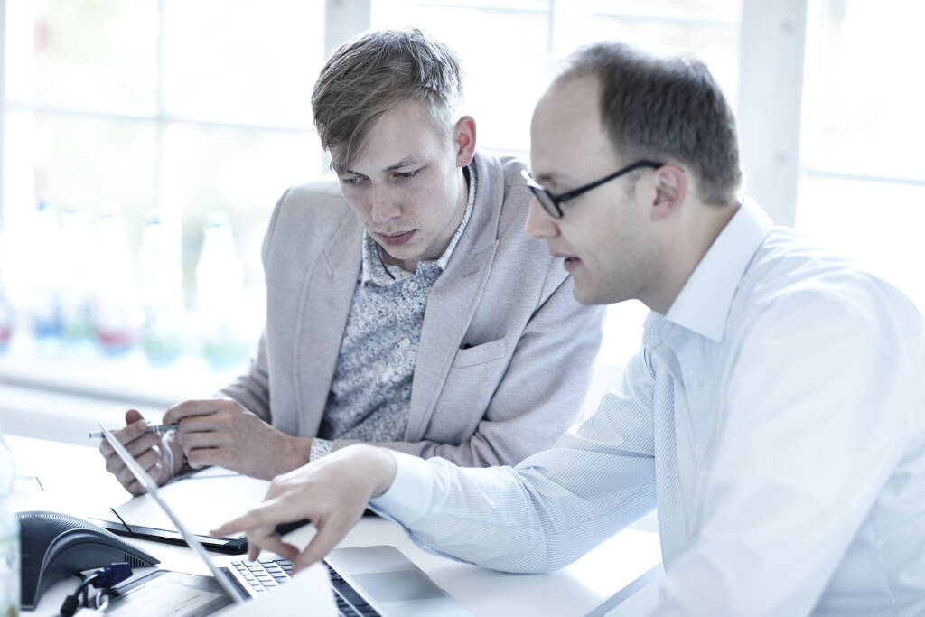 Zwei junge, hellhäutige Männer in einem hellen Büro beugen sich über einen Laptop; der Mann mit Brille in einem hellblauen Hemd zeigt auf den Bildschirm, während der andere Mann im beigefarbenen Sakko aufmerksam zusieht.