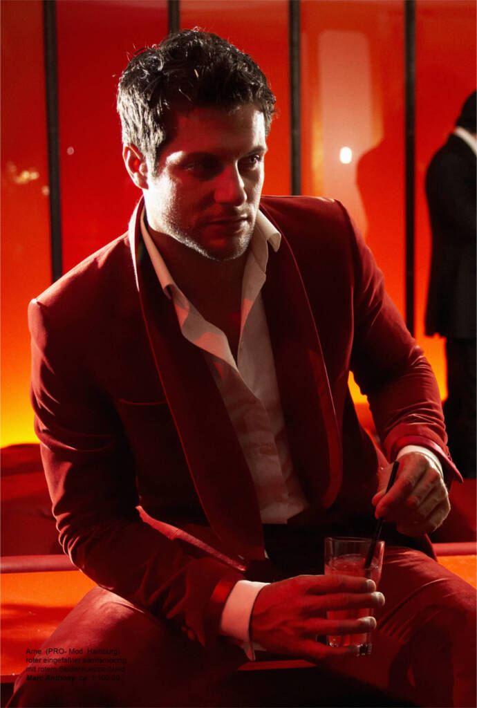 Ein stilvoller Mann in einem dunkelroten Samt-Smoking und einem offenen weißen Hemd sitzt in einem Club mit roter Beleuchtung, hält ein Cocktailglas in der Hand und blickt nachdenklich.