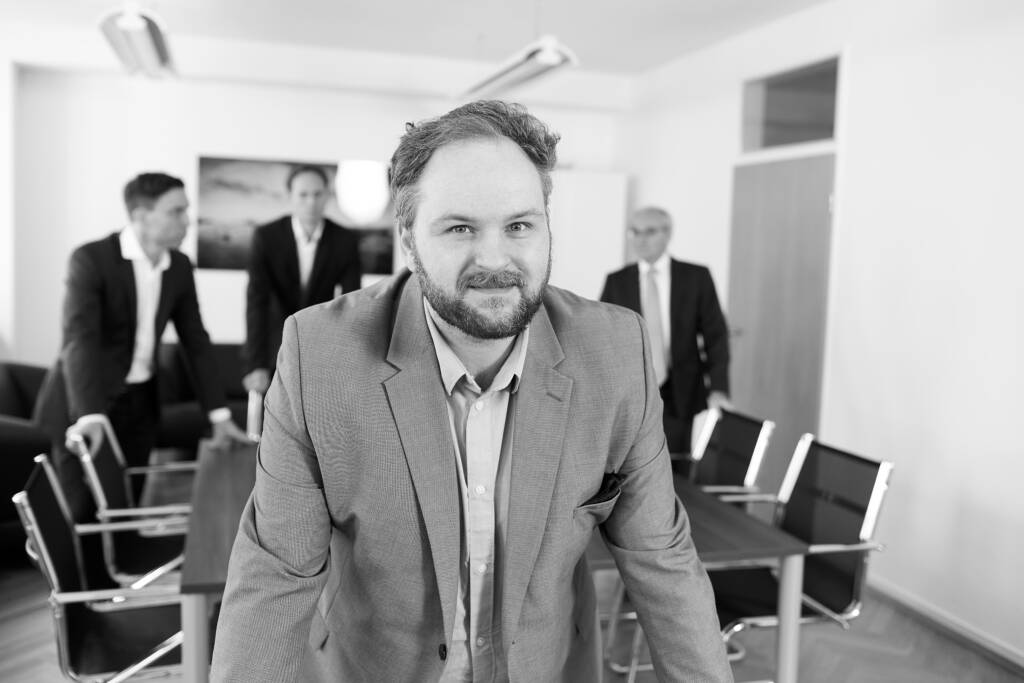 Schwarz-Weiß-Porträt eines bärtigen Mannes in einem Sakko, der sich lächelnd über einen Konferenztisch beugt, während im unscharfen Hintergrund drei weitere Männer in Anzügen im Besprechungsraum stehen.